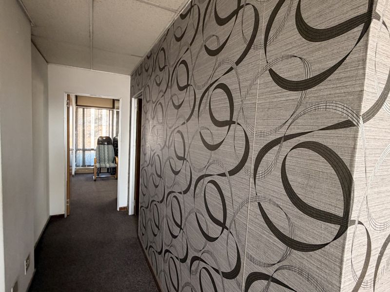 A narrow office hallway featuring patterned wallpaper, carpeted flooring, and a drop ceiling, leading into an adjacent office space.