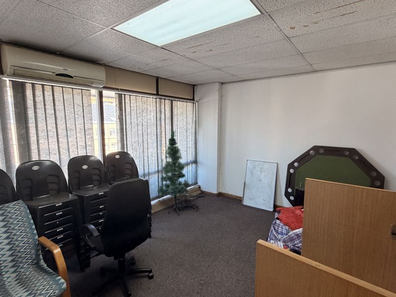 A functional office space featuring a drop ceiling with recessed lighting, wall-mounted air conditioning, and large windows with vertical blinds.