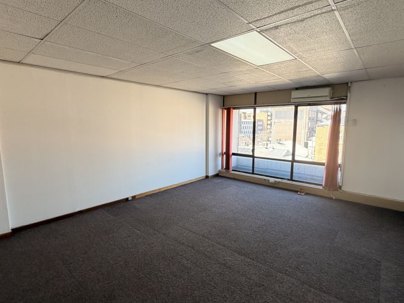 An empty office space featuring a suspended ceiling, large window with natural light, and a wall-mounted air conditioning unit.