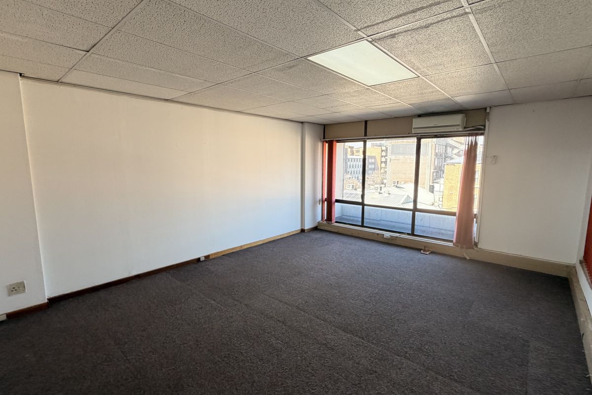 An empty office space featuring a suspended ceiling, large window with natural light, and a wall-mounted air conditioning unit.