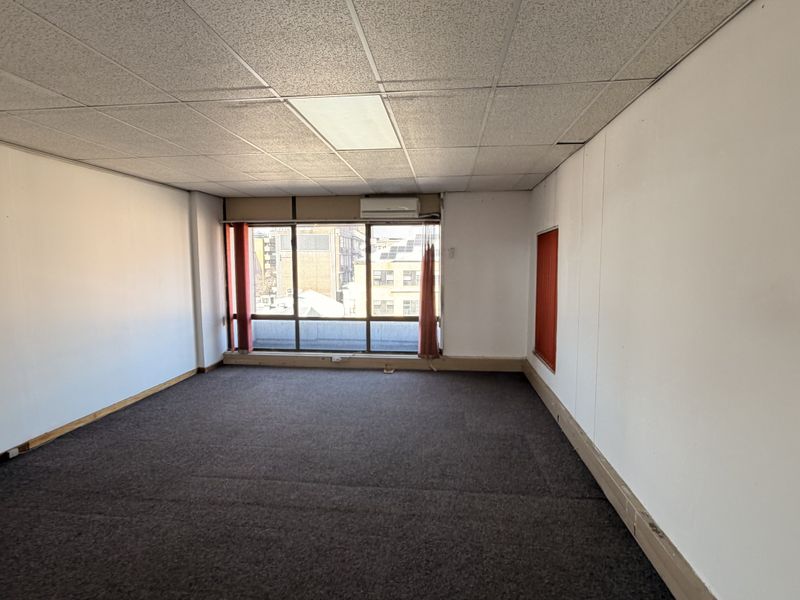 An empty, open-plan office space featuring a suspended ceiling, recessed lighting, split-system air conditioning, and large windows providing natural light.