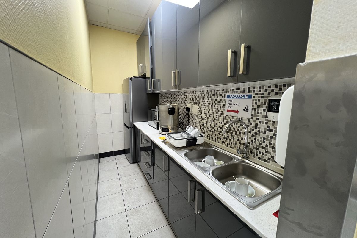 A compact office kitchenette featuring modern dark cabinetry, a double stainless steel sink, mosaic tile backsplash, and tiled flooring.