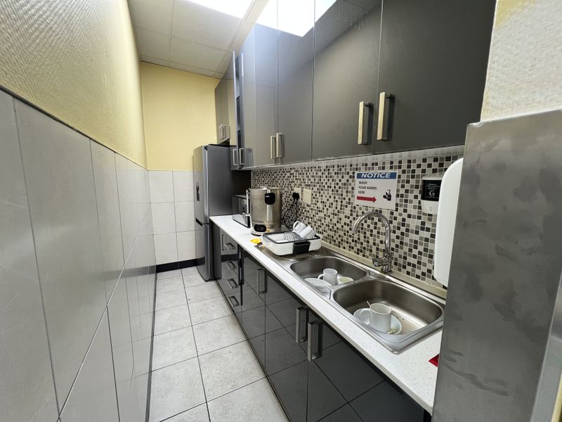 A compact office kitchenette featuring modern dark cabinetry, a double stainless steel sink, mosaic tile backsplash, and tiled flooring.