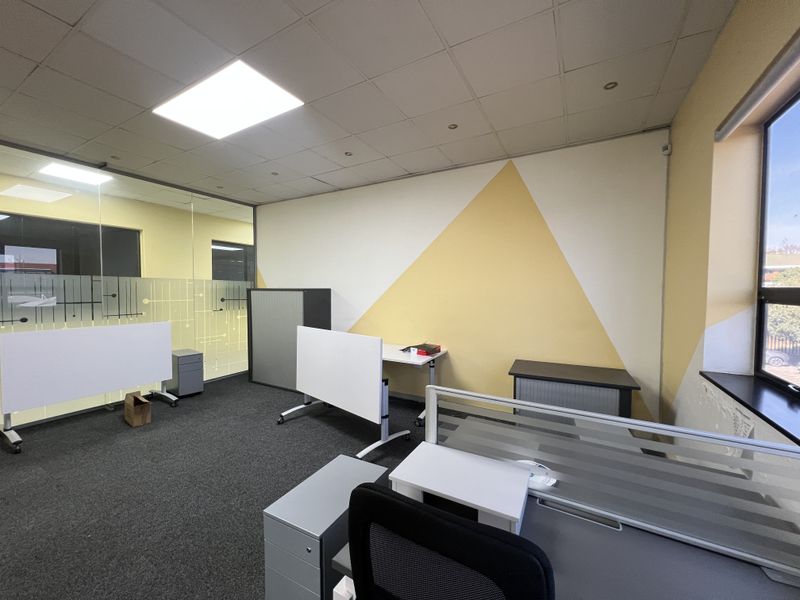 A functional office space featuring a glass partition, suspended ceiling with recessed lighting, and large windows providing natural light.
