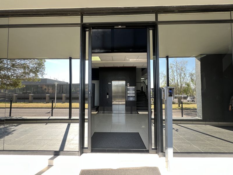 Modern office building entrance featuring a glass facade, secure intercom access, and a central elevator lobby with tiled flooring.