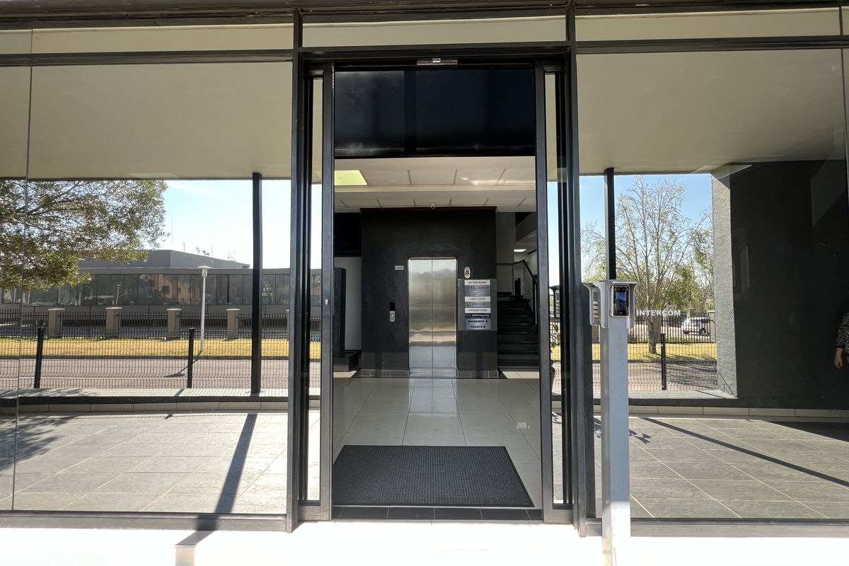 Modern office building entrance featuring a glass facade, secure intercom access, and a central elevator lobby with tiled flooring.