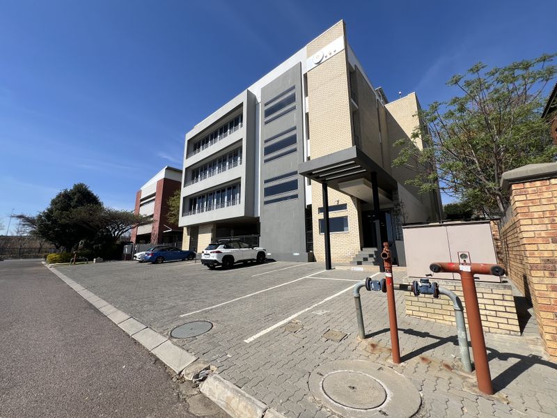 Modern multi-story office building featuring a paved parking area, covered entrance, and brick and concrete facade with large windows.