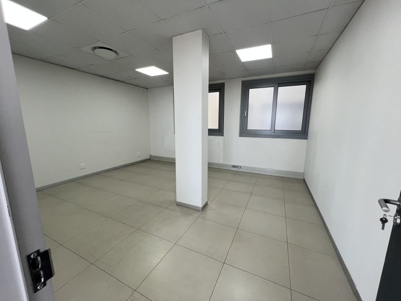 A clean, vacant office space featuring neutral tiled flooring, a suspended ceiling with recessed lighting, and a central structural column.