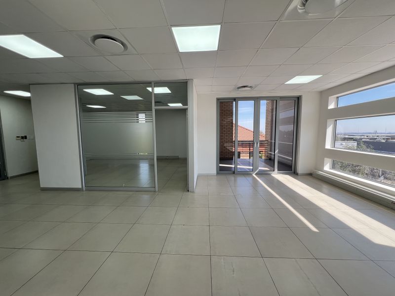 A bright, open-plan office space featuring tiled flooring, a glass-partitioned meeting room, and large windows with access to a balcony.
