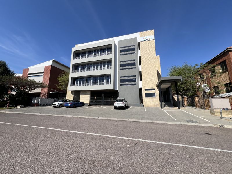 A modern multi-story office building featuring a paved front parking area, secure gated access, and a covered entrance canopy.