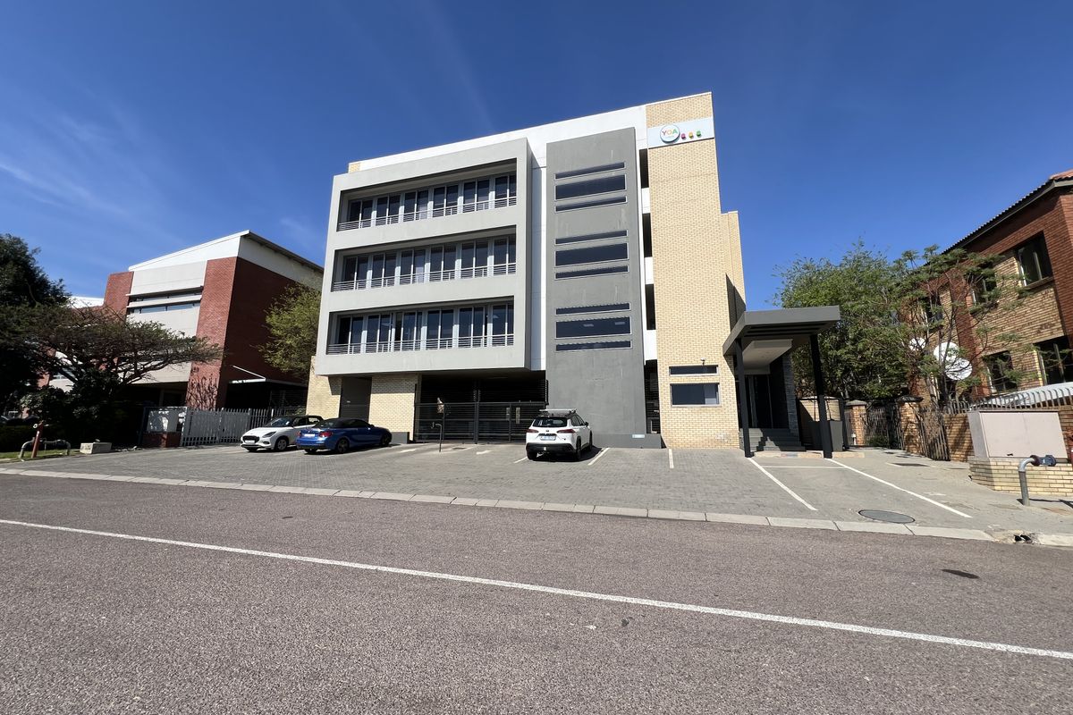A modern multi-story office building featuring a paved front parking area, secure gated access, and a covered entrance canopy.