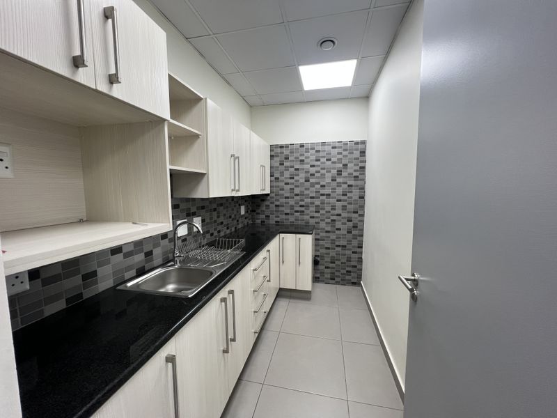 A clean and modern kitchenette featuring light wood-grain cabinetry, black countertops, a stainless steel sink, and a decorative mosaic tile backsplash.