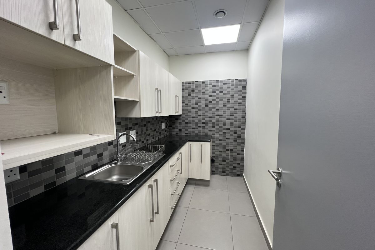 A clean and modern kitchenette featuring light wood-grain cabinetry, black countertops, a stainless steel sink, and a decorative mosaic tile backsplash.