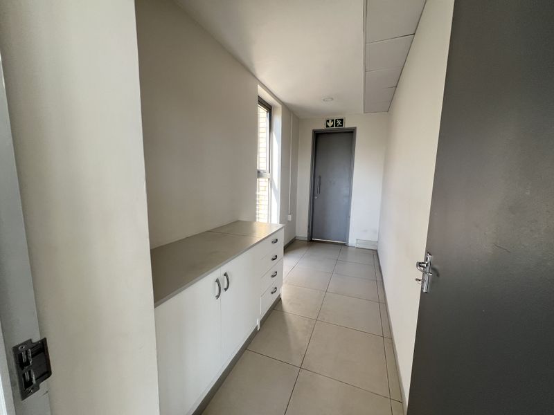 A clean, functional office hallway featuring neutral tiled flooring, built-in storage cabinetry, and a designated fire exit door.