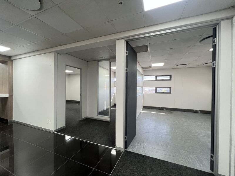 A bright, modern office space featuring a mix of carpet and vinyl flooring, glass partitions, and a suspended ceiling with recessed lighting.