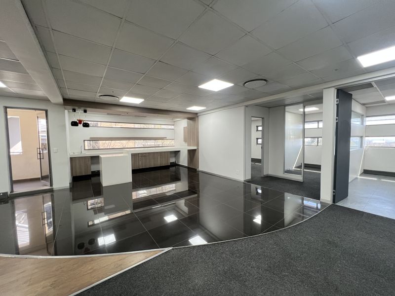 Modern open-plan office space featuring a built-in kitchenette, glass partitions, suspended ceilings with recessed lighting, and a combination of tiled and carpeted flooring.