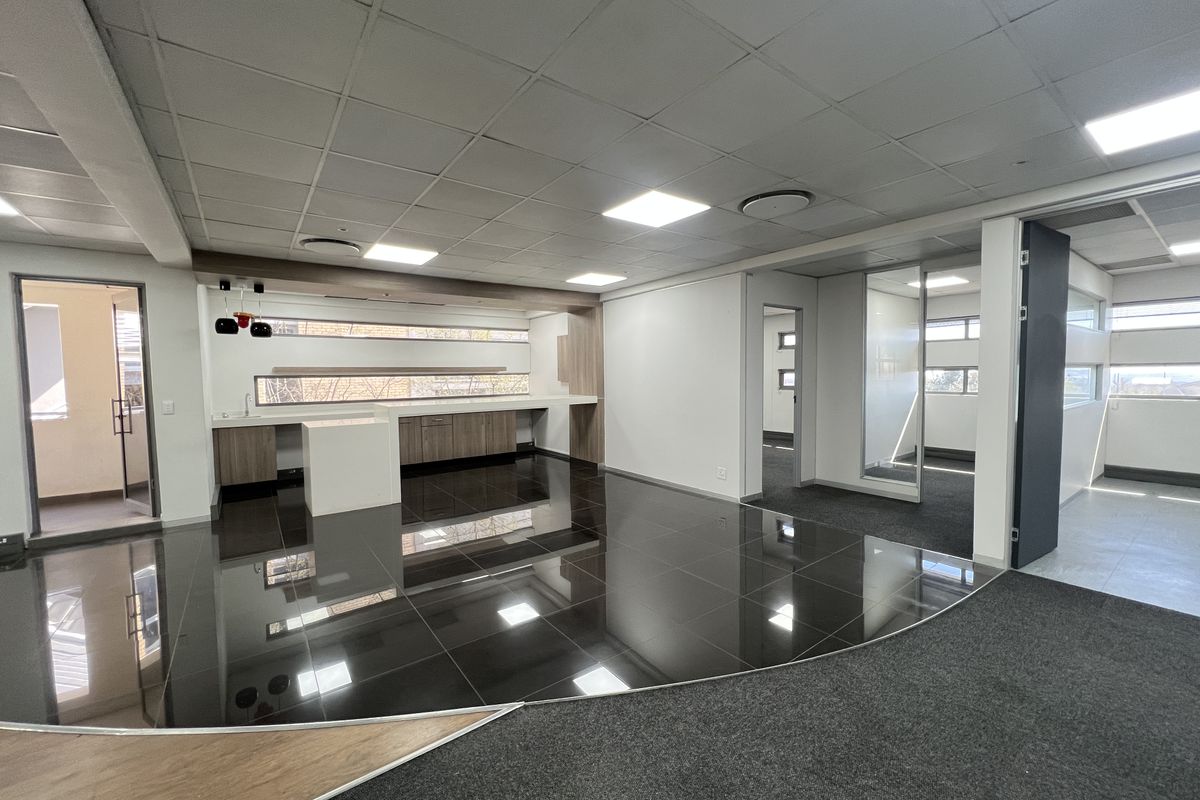 Modern open-plan office space featuring a built-in kitchenette, glass partitions, suspended ceilings with recessed lighting, and a combination of tiled and carpeted flooring.