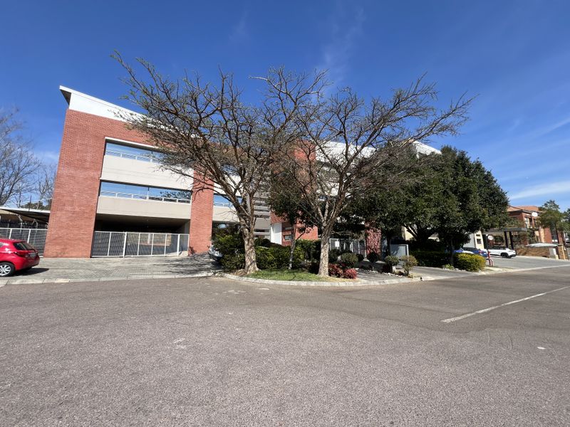 Front exterior view of a multi-story office building featuring a red brick facade, paved parking area, and established landscaping.