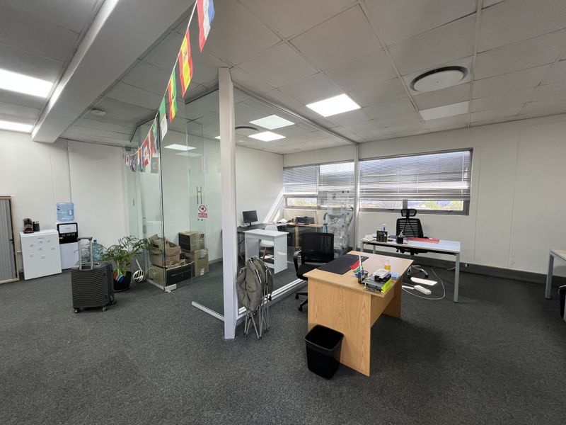 Open-plan office space featuring a glass-partitioned private office, suspended ceiling with recessed lighting, and large windows with venetian blinds.
