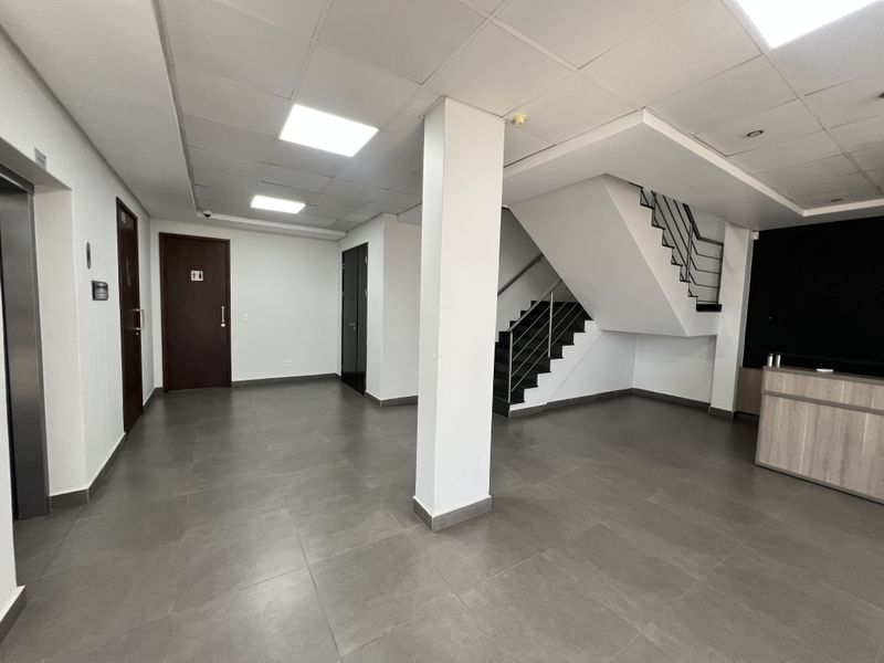 Spacious office lobby featuring tiled flooring, a central staircase with metal railings, elevator access, and a built-in reception desk.