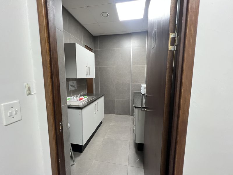 A compact office kitchenette featuring modern grey wall tiles, white cabinetry, laminate countertops, and a stainless steel sink.