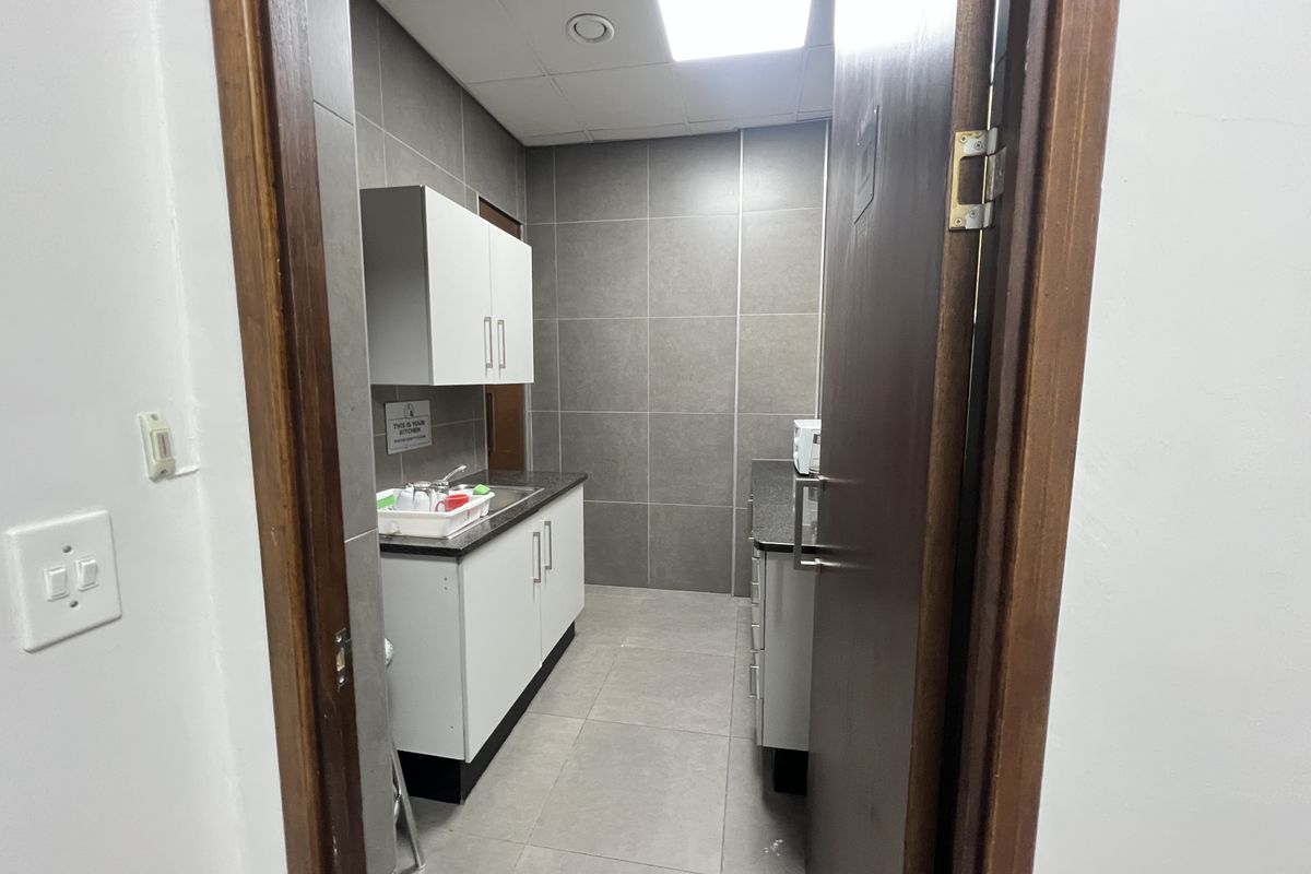 A compact office kitchenette featuring modern grey wall tiles, white cabinetry, laminate countertops, and a stainless steel sink.