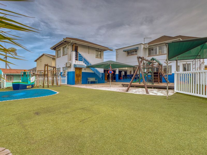 Rear exterior view of a commercial property featuring a large artificial turf play area, paved patio, shade sails, and external staircase access.
