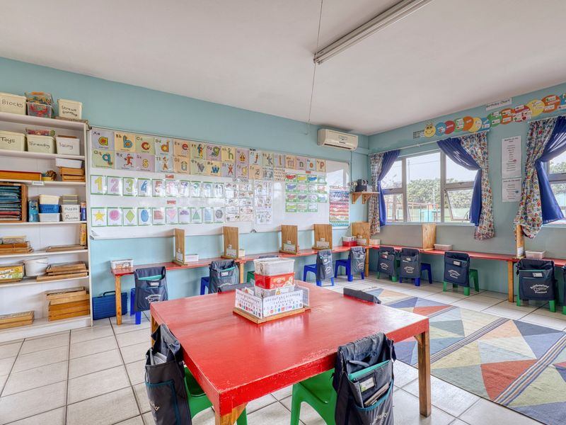 A functional classroom space featuring tiled flooring, built-in storage shelving, an air conditioning unit, and large windows providing natural light.