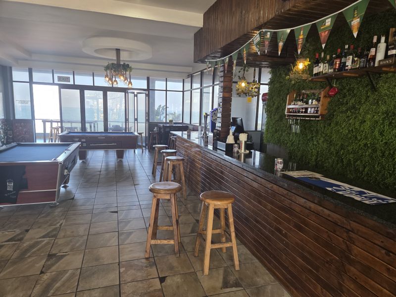 A spacious restaurant bar area featuring a long wooden counter, tiled flooring, and large glass doors leading to an outdoor space.