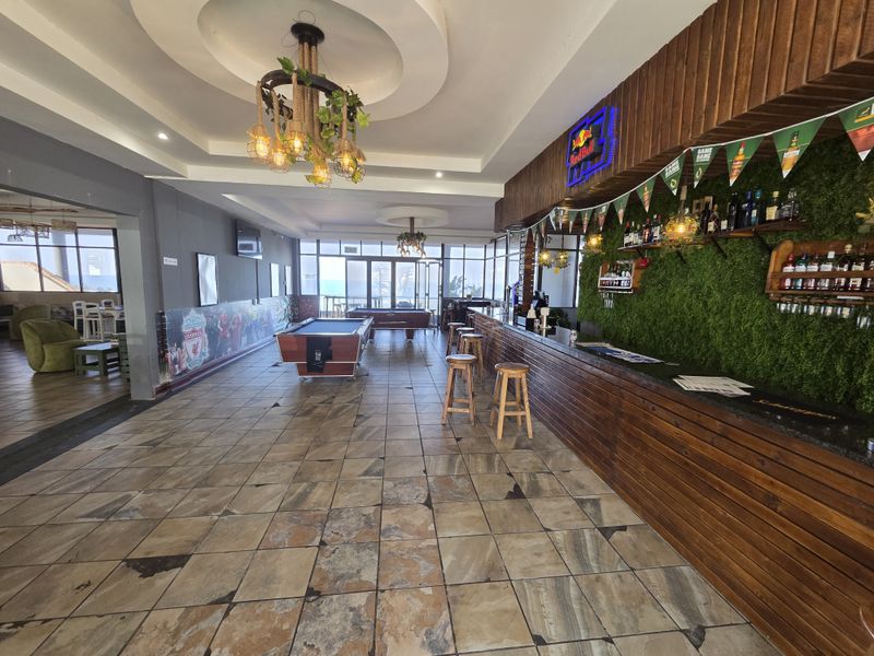 Spacious restaurant bar area featuring a long built-in counter, decorative green wall, tiled flooring, and recessed ceiling lighting with views toward the exterior.