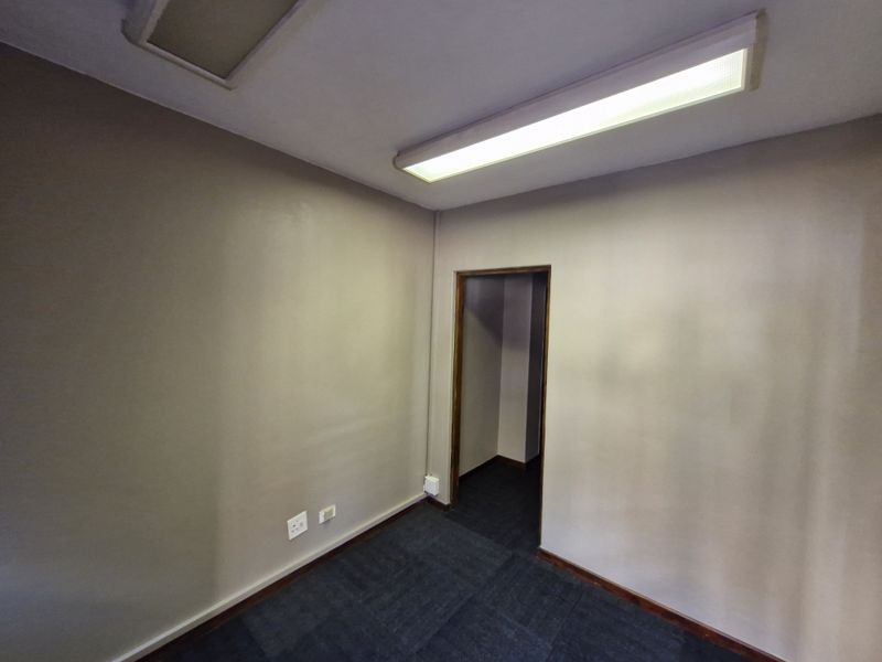 A vacant office space featuring neutral wall finishes, dark carpet tiles, and a functional overhead fluorescent lighting fixture.