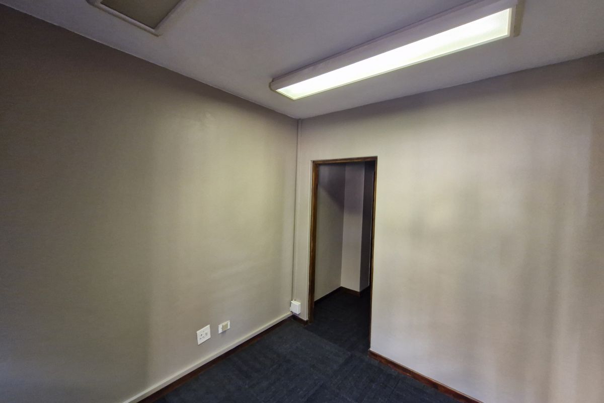 A vacant office space featuring neutral wall finishes, dark carpet tiles, and a functional overhead fluorescent lighting fixture.