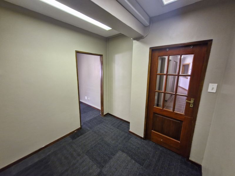 A vacant office space featuring carpet tile flooring, fluorescent lighting, and a classic timber-framed glass door leading to an adjacent hallway.