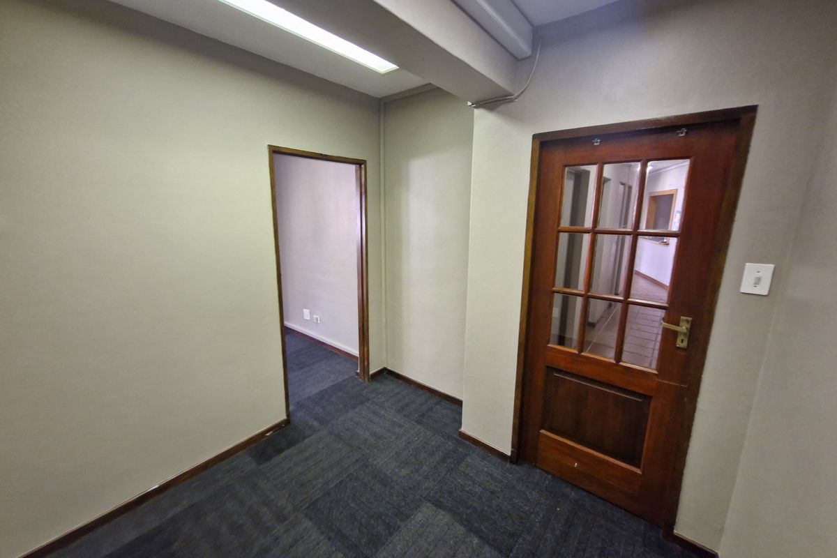 A vacant office space featuring carpet tile flooring, fluorescent lighting, and a classic timber-framed glass door leading to an adjacent hallway.