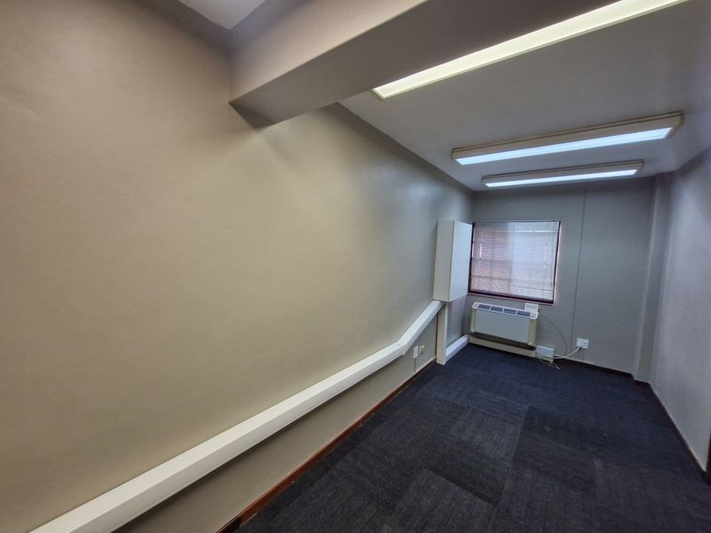 A vacant office space featuring carpet tiles, surface-mounted electrical trunking, fluorescent lighting, and a wall-mounted air conditioning unit.