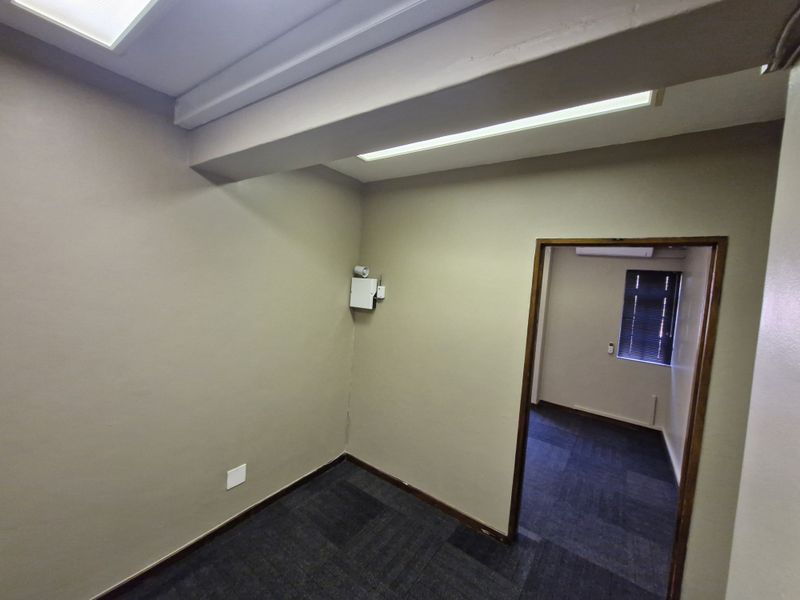 A vacant office space featuring carpeted flooring, fluorescent lighting, and an integrated air conditioning unit, with a view into an adjacent office area.