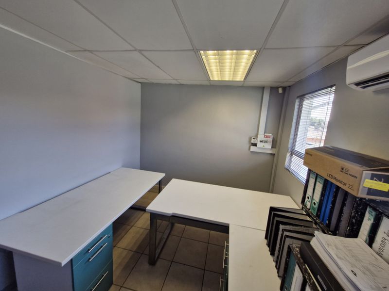 A functional office space featuring tiled flooring, a suspended ceiling with recessed lighting, and a wall-mounted air conditioning unit.