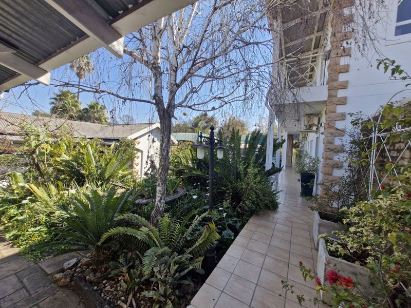 A view of a rear garden area featuring a tiled walkway, stone-clad building facade, and established landscaping with a covered patio section.