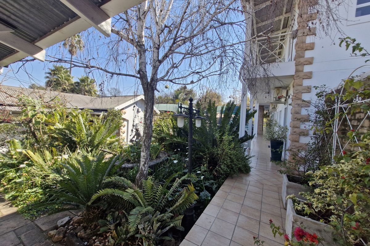A view of a rear garden area featuring a tiled walkway, stone-clad building facade, and established landscaping with a covered patio section.