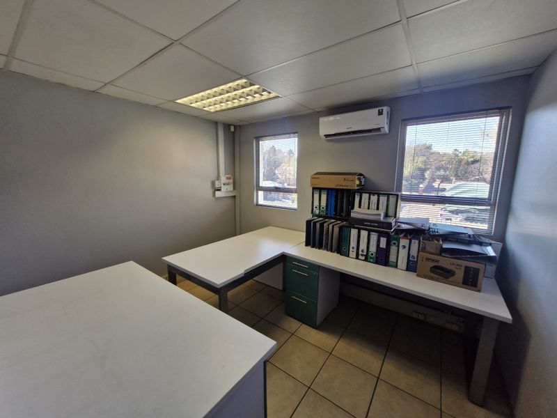 A functional office space featuring tiled flooring, a suspended ceiling with recessed lighting, and a wall-mounted air conditioning unit.