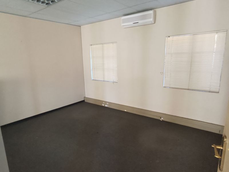 A vacant office space featuring carpeted flooring, a suspended ceiling, wall-mounted air conditioning, and windows fitted with venetian blinds.