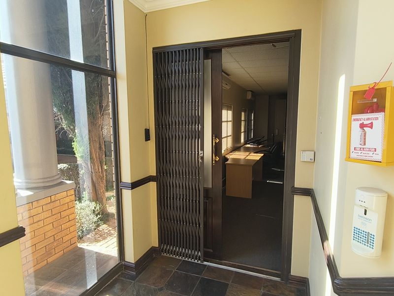 A professional office entrance area featuring a security gate, emergency alarm station, intercom system, and tiled flooring with access to an office space.
