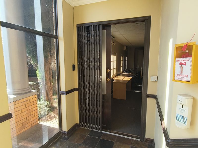 A bright office entrance area featuring tiled flooring, a security gate, and wall-mounted fire safety equipment, leading into a professional workspace.