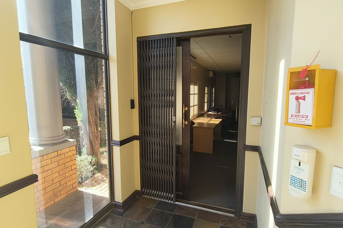 A bright office entrance area featuring tiled flooring, a security gate, and wall-mounted fire safety equipment, leading into a professional workspace.