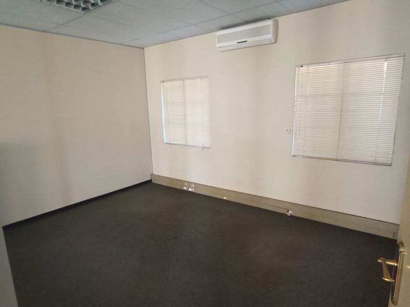 A vacant office space featuring carpeted flooring, a suspended ceiling, wall-mounted air conditioning, and windows fitted with venetian blinds.