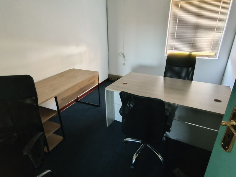 A functional office space featuring carpeted flooring, a window with venetian blinds, and a standard door entrance.
