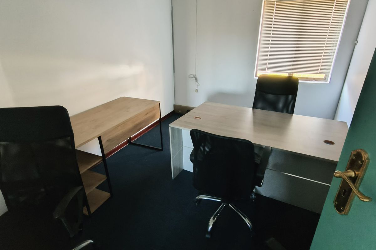A functional office space featuring carpeted flooring, a window with venetian blinds, and a standard door entrance.