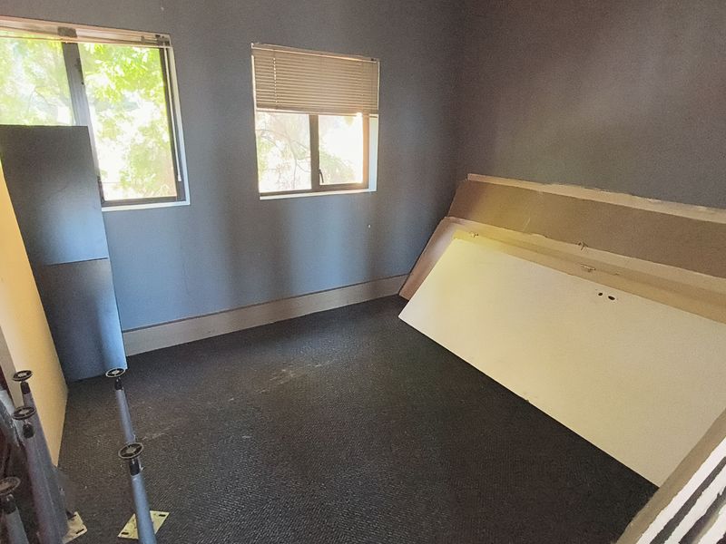 A vacant office space featuring carpeted flooring, dual windows with venetian blinds, and grey painted walls, currently undergoing minor maintenance or renovation.