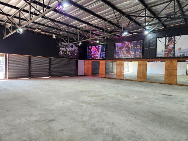 Spacious industrial-style gym area featuring exposed steel roof trusses, concrete flooring, mirrored walls, and roller shutter doors, suitable for a fitness facility.