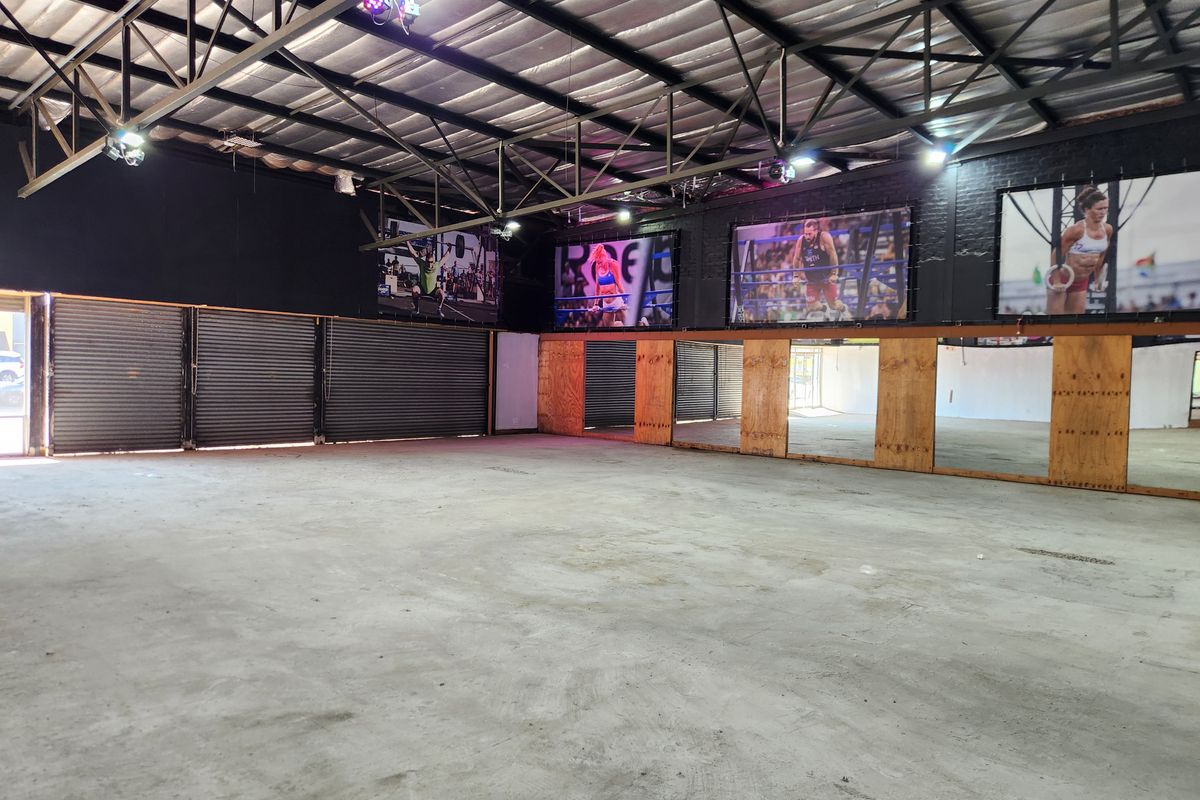 Spacious industrial-style gym area featuring exposed steel roof trusses, concrete flooring, mirrored walls, and roller shutter doors, suitable for a fitness facility.